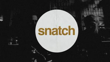 A large white circle with the word "snatch" in lowercase letters on top of a dark image of two men sitting in a pub