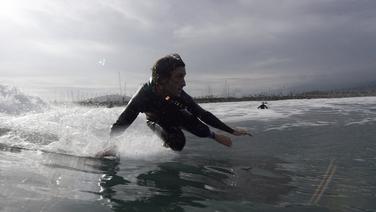 A man in a wetsuit surfing a small wave with an industrial factory in the background.