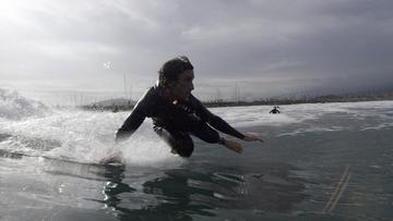 A man in a wetsuit surfing a small wave with an industrial factory in the background.