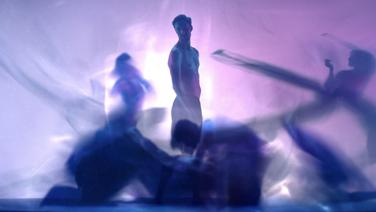 A shirtless young man standing amongst out-of-focus figures in flowy clothing.