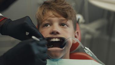 A young boy in a dentist's chair with mouth held open. On one side gloved hands are cleaning his teeth and on the other the saliva sucker is inserted.