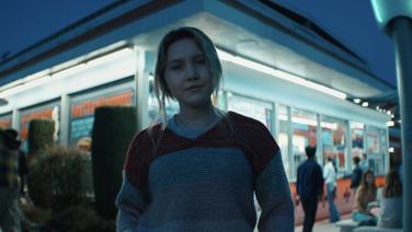 A young woman standing in the outdoor seating area of a restaurant at dusk.