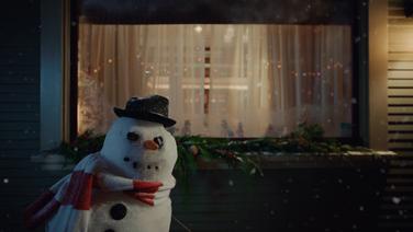 A snowman wearing a black pork pie hat and a red and white striped scarf standing at a lean in front of a living room window draped with white sheer curtains.