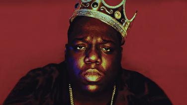 Biggie Smalls wearing a crown and a gold necklace looking straight ahead.