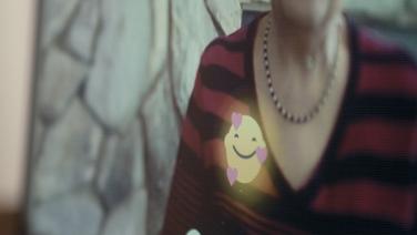 A slightly pixelated video still of a young white girl wearing a red and black striped shirt with a smiley face emoji and three hearts around it.