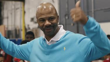 A bald middle-aged black man wearing an aqua blue sweater with a white collared shirt beneath. His arms are raised with both thumbs up.