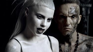 A pale white woman with long blond hair with very short bangs wearing dark lipstick stands next to a man with mud all over his face and body.
