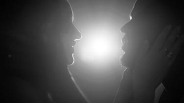 A man and a woman holding each other's faces with a bright light between them.