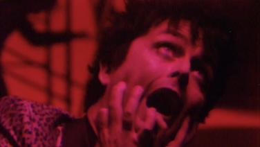 A red-tinted image of Bille Joe Armstrong of Green Day clutching his face with mouth agape.