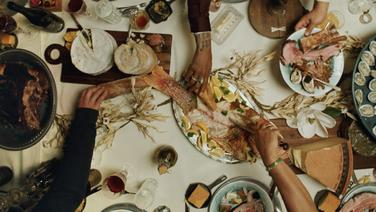 Three hands reaching toward a table full of various food dishes seen from above.