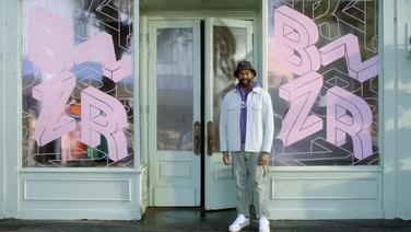 A middle-aged black man with a beard standing in front of a pale green storefront with the door ajar.