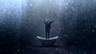 A clothed man standing in a bathtub with arms raised and head up with rain pouring down all aroud him.
