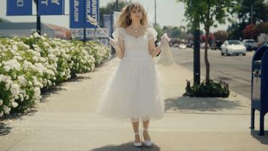 A middle-aged woman dressed as a fairy in a white gown holding a wand and a white bag standing on a sidewalk in a suburban business area.